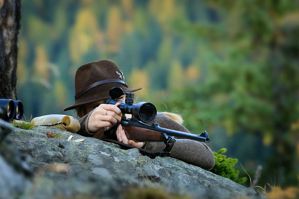 Joven y astuta cazadora de ciervos con uniforme verde y un rifle, sentada sobre una roca con árboles otoñales al fondo en Trofeo caza y conservacion